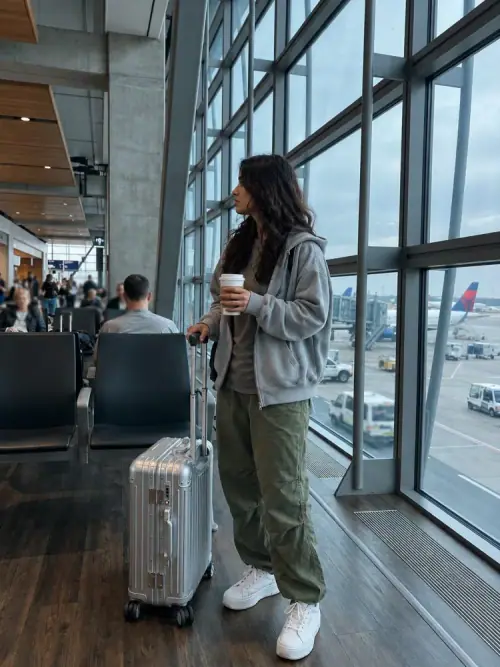 Airport Travel Outfit AI Photo Prompt showing a stylish Indian woman in a grey hoodie, olive cargo trousers, and white sneakers holding coffee beside a silver suitcase at an airport window.
