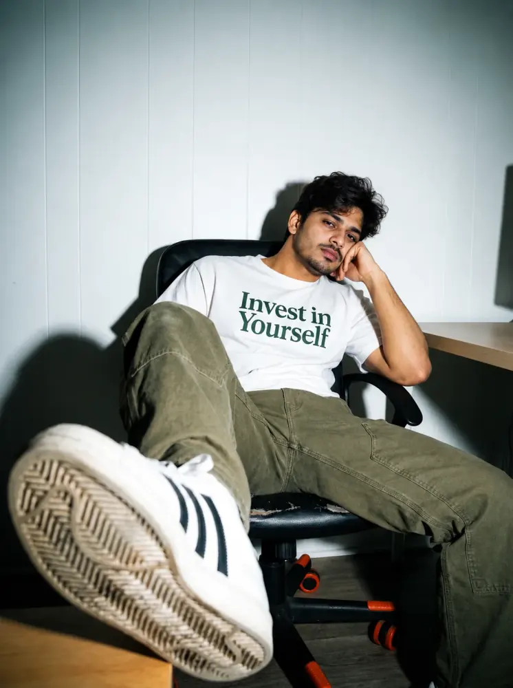 Indian Man Sitting Portrait AI Photo Prompt showing a young Indian man lounging in a black office chair, wearing a white graphic T-shirt, olive cargo pants, and white sneakers.